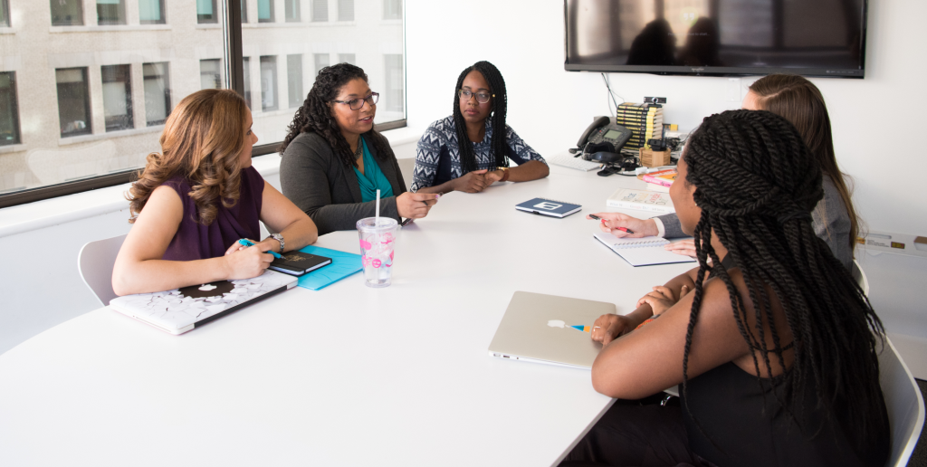 Picture of 5 women sitting around a conference room table discussing something at a meeting.